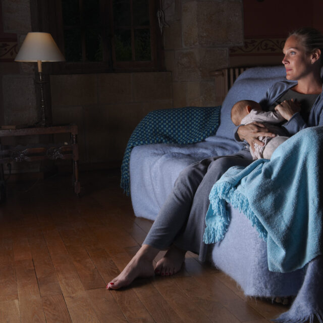 A woman breastfeeds a baby on the couch while watching television