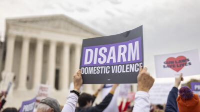 A sign is held up in front of the Supreme Court. Sign has a black background and reads 