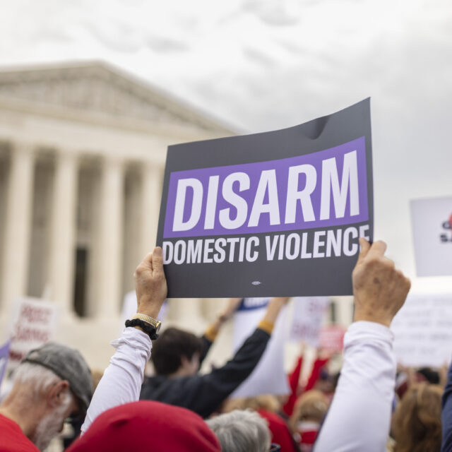 A sign is held up in front of the Supreme Court. Sign has a black background and reads