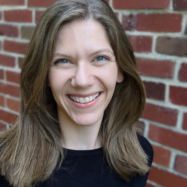 A white woman with shoulder-length brown hair and blue eyes is pictured from the shoulders up. She wears a black shirt and is smiling. A brick wall is the background behind her.