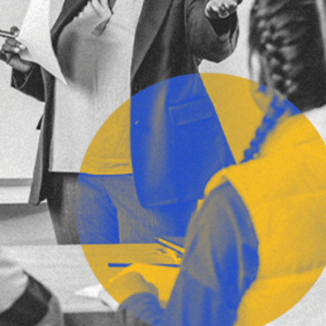 A black-and-white photo of two students sitting at a desk, looking at a teacher who is standing at a whiteboard. The photo is overlaid with a yellow and blue circle.