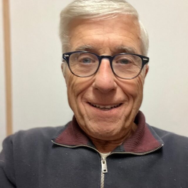 Dr. Archie Bleyer takes a selfie. He is standing in front of a blank white wall; he wears cat-eye glasses with a black frame and a zip-up maroon, navy, and white sweater.