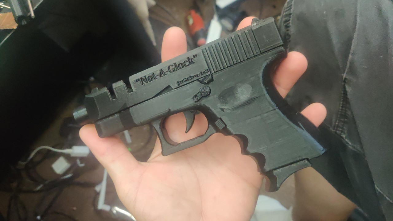 A person holds a black 3d-printed "Not-A-Glock" gun in the palm of their left hand; it is lying flat on their hand so the text is easier to read. "Not-A-Glock" purportedly copies the Glock 26.